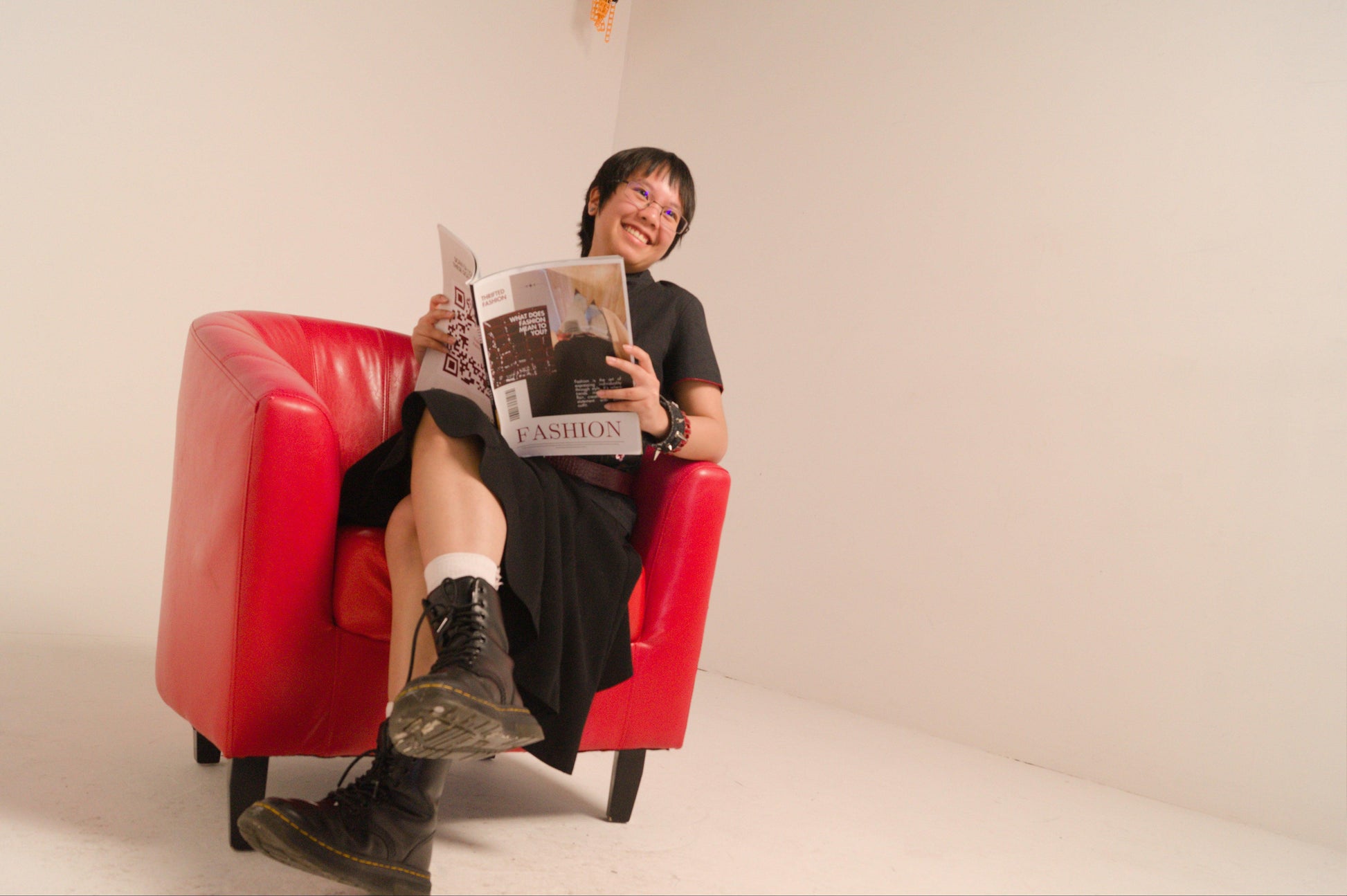 Smiling person in a black flowy midi skirt, black top, and boots, sitting in a red armchair, reading a fashion magazine.