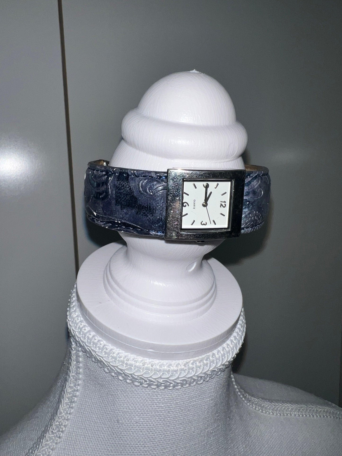 Silver square face watch with a blue patterned pleather strap on a white display.